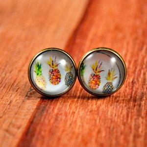 May include: A pair of gold-toned stud earrings with a clear glass cabochon featuring a watercolor-style design of pineapples.