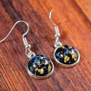 May include: A pair of silver dangle earrings with black round resin cabochons. The cabochons are filled with gold and blue glitter.