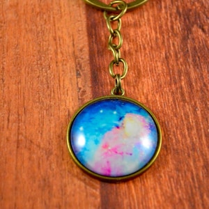 May include: A galaxy-themed keychain with a round glass charm featuring a blue, pink, and white galaxy design. The charm is attached to a gold-colored chain and a key ring.