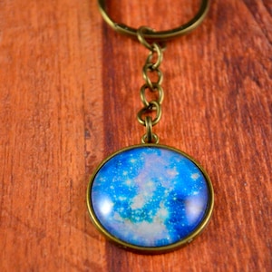 May include: A galaxy-themed keychain with a blue, white, and pink galaxy design inside a round glass charm. The charm is attached to a gold-colored chain and ring.