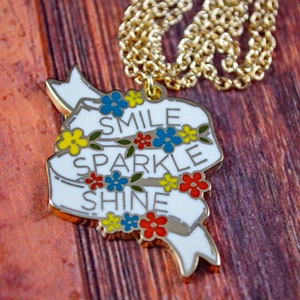 May include: A gold chain necklace with a white enamel pendant that says "Smile Sparkle Shine" with colorful flowers around the words.