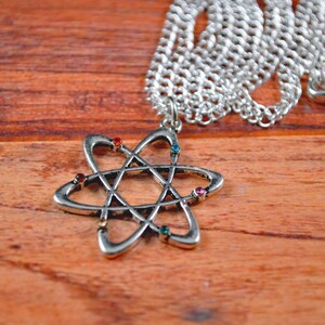 May include: A silver necklace with a silver atom pendant featuring colorful rhinestones. The necklace is on a wooden surface.