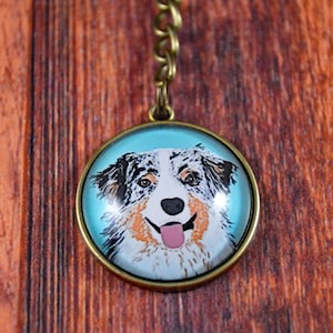 May include: A round glass keychain with a blue background and a picture of an Australian Shepherd dog with brown, black, and white fur. The dog is looking at the camera and has its tongue sticking out.