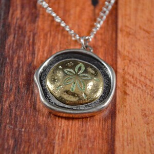 May include: A silver necklace with a pendant. The pendant has a gold-coloured sand dollar design with green detailing, set in a textured, silver-coloured frame. The necklace is displayed on a wooden surface.