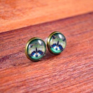 May include: A pair of small, round earrings with a green background and a raccoon face image. The earrings have a gold-colored metal setting.