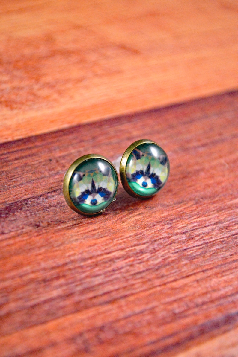 May include: A pair of small, round earrings with a green background and a raccoon face image. The earrings have a gold-colored metal setting.