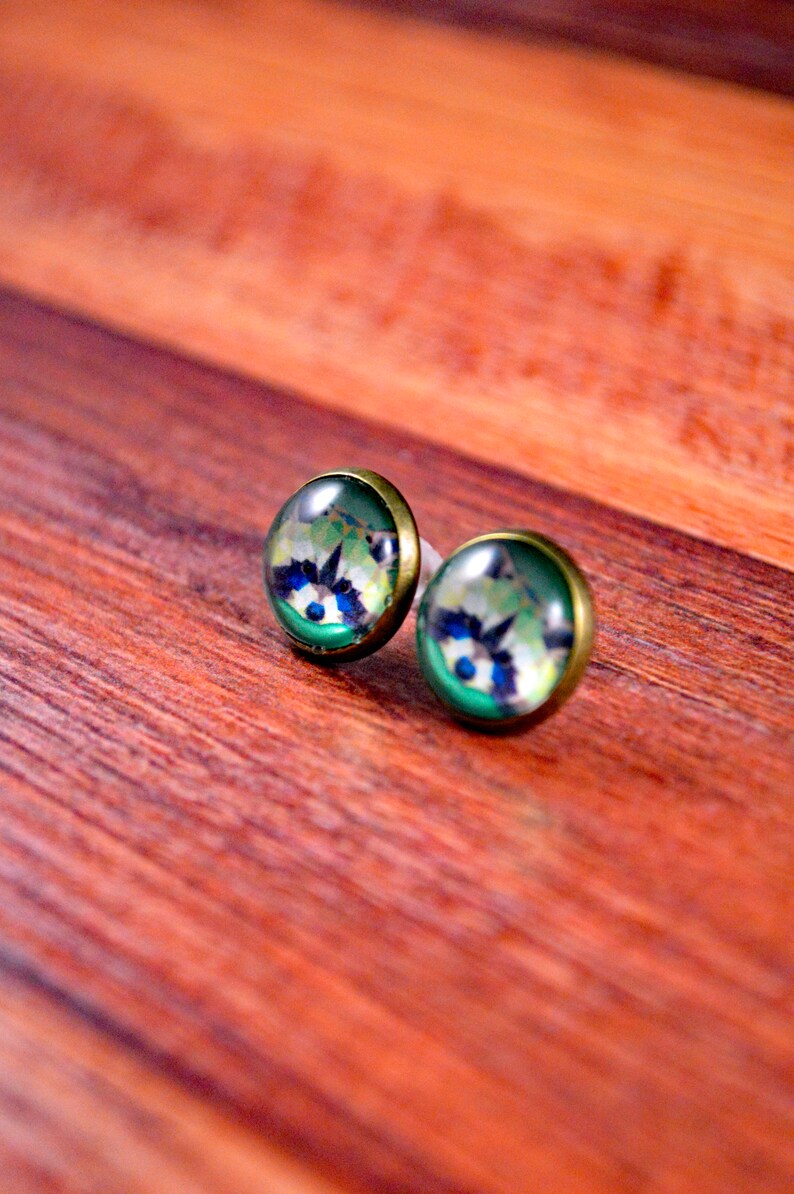May include: A pair of stud earrings with a green, blue, and white raccoon design. The earrings are made of metal and have a shiny finish.