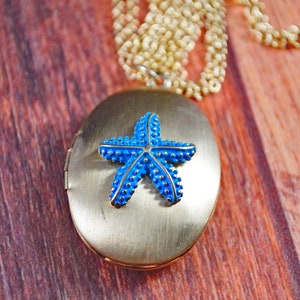 May include: A gold locket necklace with a blue enamel starfish charm. The locket is oval shaped and has a hinged lid.