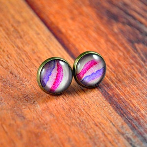 May include: A pair of stud earrings with a pink, purple, and white abstract design. The earrings have a bronze-colored metal setting.