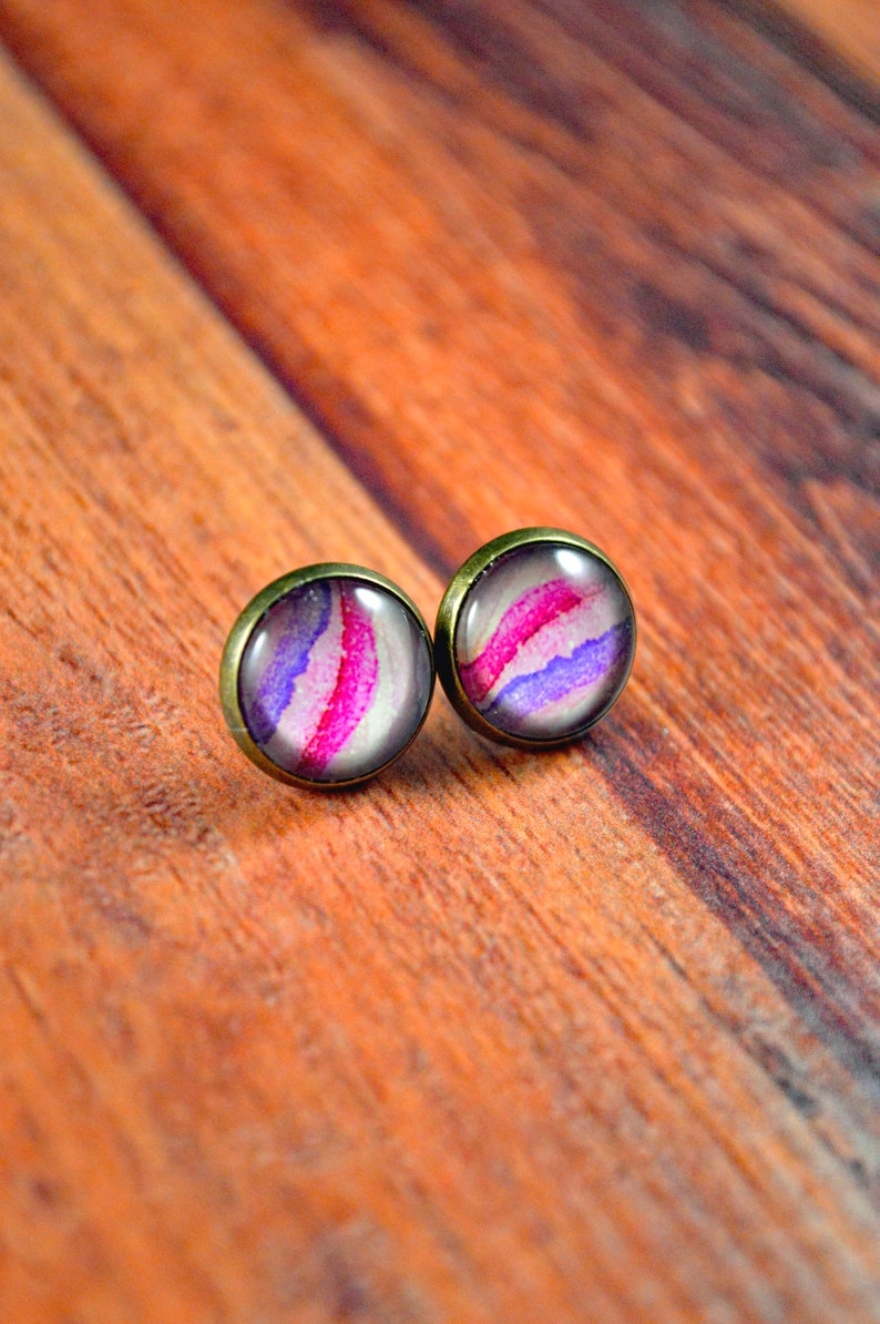 May include: A pair of stud earrings with a pink, purple, and white abstract design. The earrings have a bronze-colored metal setting.