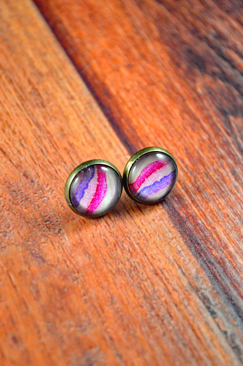 May include: A pair of stud earrings with a pink, purple, and white abstract design. The earrings have a bronze-colored setting.
