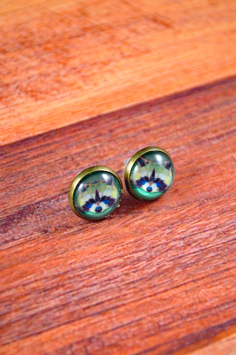 May include: A pair of bronze-colored stud earrings with a green, blue, and white raccoon face design.