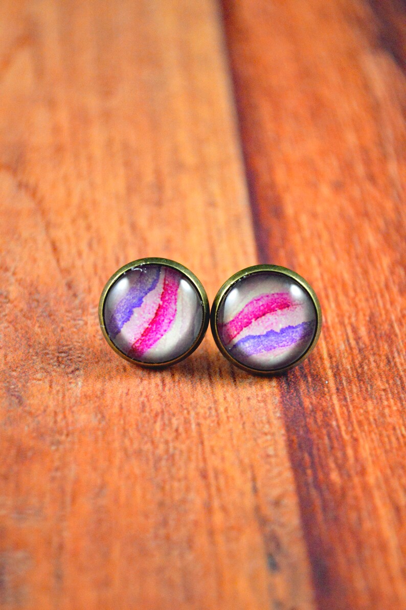 May include: A pair of stud earrings with a purple, pink, and white abstract design. The earrings have a bronze-colored metal setting.