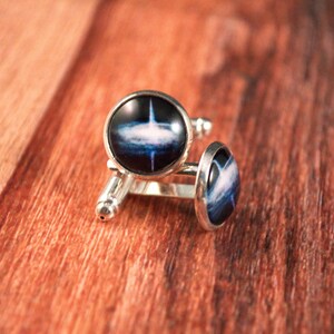 May include: A pair of silver cufflinks with a blue and white galaxy design. The cufflinks are on a wooden surface.