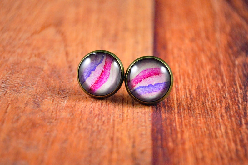 May include: A pair of stud earrings with a round, white background and a pink and purple watercolor design.