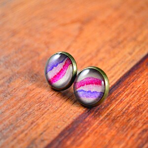 May include: A pair of small, round earrings with a bronze setting. The earrings feature a white background with pink and purple watercolor stripes.