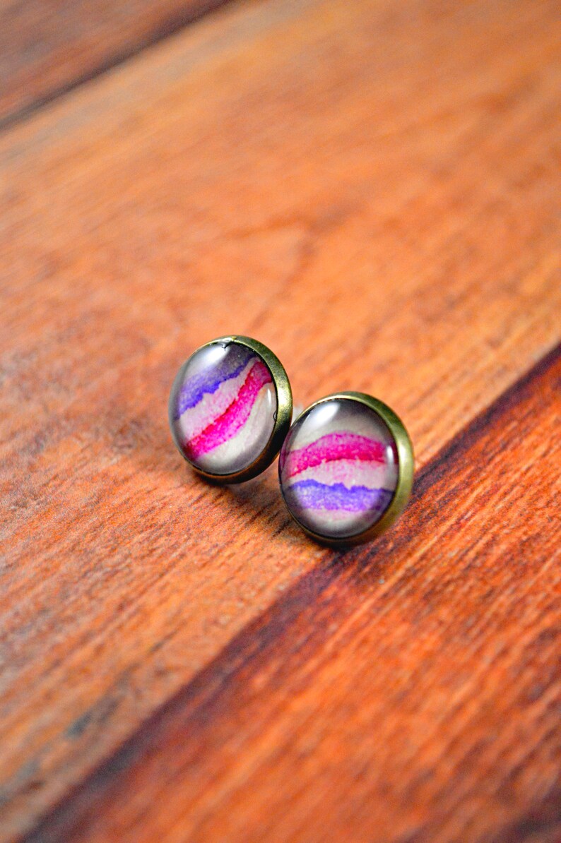 May include: A pair of small, round earrings with a bronze setting. The earrings feature a white background with pink and purple watercolor stripes.