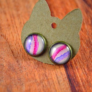 May include: A pair of stud earrings with a pink and purple watercolor design. The earrings are on a brown card with a hole in the center.