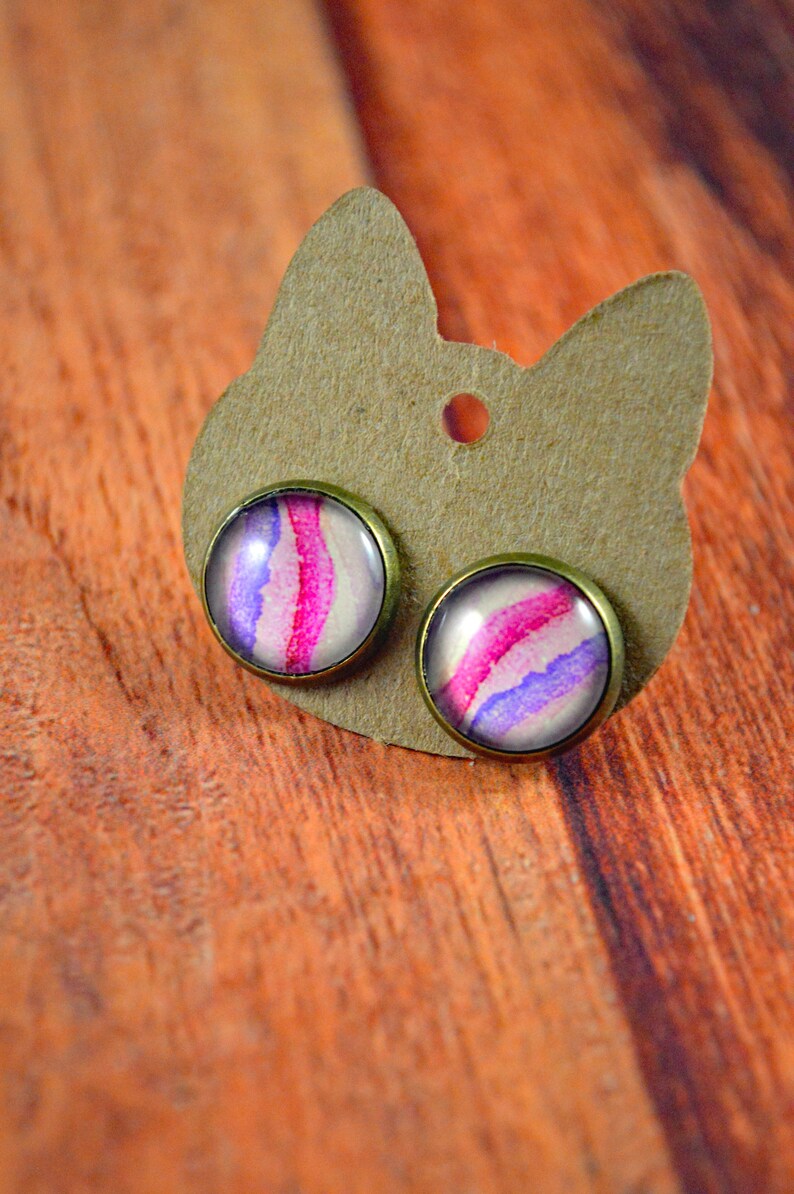 May include: A pair of stud earrings with a pink and purple watercolor design. The earrings are on a brown card with a hole in the center.