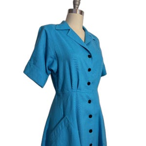 May include: A light blue short-sleeved dress with a collared neckline and a button-up front. The dress has a fitted bodice and a flared skirt. It features two pockets on the front and black buttons.
