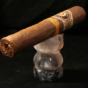 May include: A brown cigar with a gold and white band that reads "Don Carlos" is resting on a black and white marble ashtray.