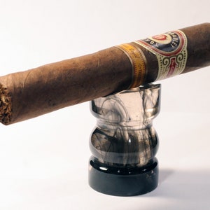 May include: A brown cigar with a gold and white band that reads "La Aurora" is resting on a black and gray glass ashtray.