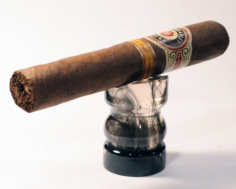 May include: A brown cigar with a gold and white band that reads "La Aurora" is resting on a black and gray glass ashtray.