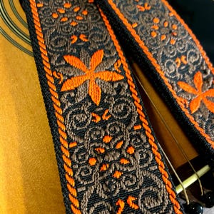 May include: Close-up of a guitar strap with a floral design. The strap features a black background with brown and orange floral patterns. The edges are trimmed with orange stitching. The strap is resting on a wooden acoustic guitar.