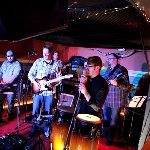 May include: A band of four musicians performing on stage. The musicians are playing a keyboard, guitar, drums, and a microphone. The stage is lit with colorful lights.