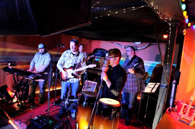 May include: A band of four musicians performing on stage. The musicians are playing a keyboard, guitar, drums, and a microphone. The stage is lit with colorful lights.