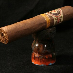 May include: A brown cigar with a gold and white label that reads "Toasted" and "Caoba" is resting on a colorful ceramic cigar holder.