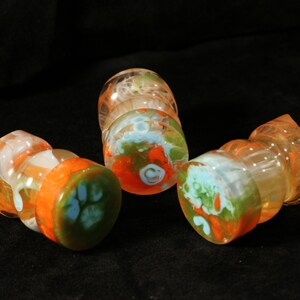 May include: Three glass bottle stoppers with a swirl design in orange, green, and white. The stoppers are made of clear glass with a swirl of orange, green, and white paint.