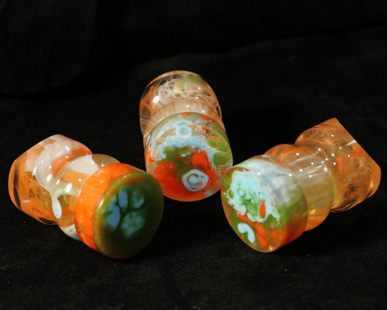 May include: Three glass bottle stoppers with a swirl design in orange, green, and white. The stoppers are made of clear glass with a swirl of orange, green, and white paint.