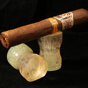 May include: A brown cigar with a gold and white band that reads "Don Francisco" is resting on two clear glass ashtrays with a white swirl design.