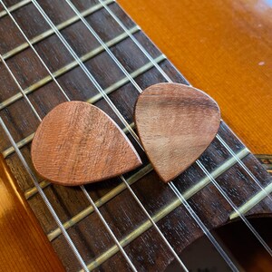 May include: Two wooden guitar picks rest on the fretboard of a classical guitar. The picks are a warm brown color, with visible wood grain. The guitar's strings and dark brown fretboard are in focus, with the guitar body in the background.