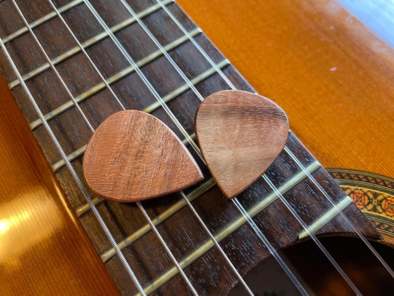 May include: Two wooden guitar picks rest on the fretboard of a classical guitar. The picks are a warm brown color, with visible wood grain. The guitar's strings and dark brown fretboard are in focus, with the guitar body in the background.