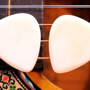 May include: Two white guitar picks, shaped like a teardrop, are placed on the strings of an acoustic guitar.