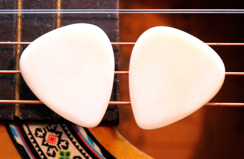May include: Two white guitar picks, shaped like a teardrop, are placed on the strings of an acoustic guitar.