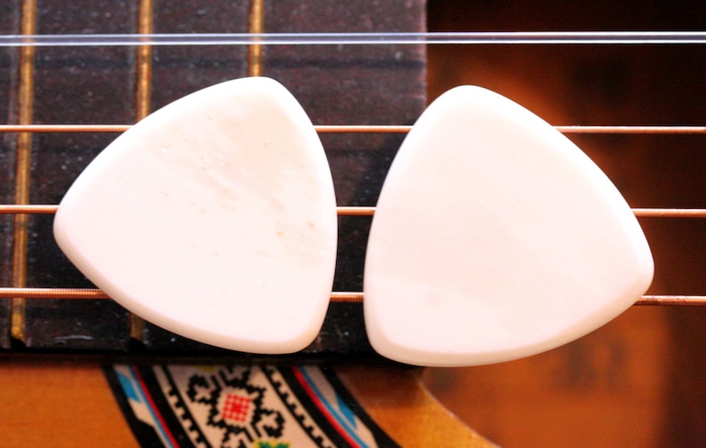 May include: Two off-white, triangular guitar picks rest on the strings of an acoustic guitar. The strings are bronze-colored, and the guitar's fretboard is dark brown. The picks are smooth and appear to be made of a solid material.