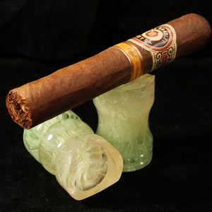 May include: A brown cigar with a white and gold label that reads "Bradley" and "Royal" is resting on two white and green decorative stands.