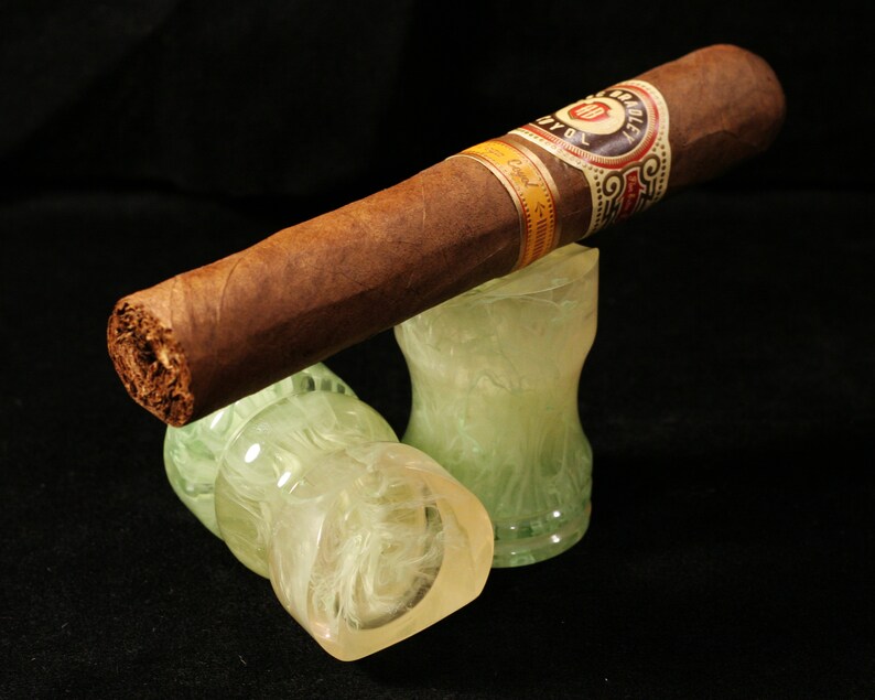May include: A brown cigar with a white and gold label that reads "Bradley" and "Royal" is resting on two white and green decorative stands.