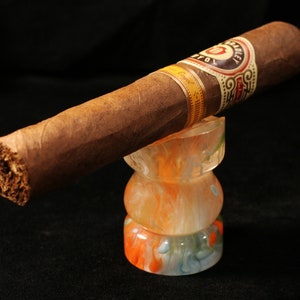 May include: A brown cigar with a gold and black band that reads "Hoyo de Monterrey" is resting on a colorful glass ashtray. The ashtray is made of clear glass with swirls of orange, white, and blue.