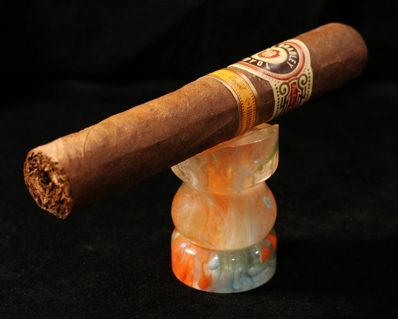 May include: A brown cigar with a gold and black band that reads "Hoyo de Monterrey" is resting on a colorful glass ashtray. The ashtray is made of clear glass with swirls of orange, white, and blue.