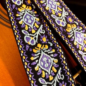 May include: Close-up of a guitar strap with a black background. The strap features a repeating pattern of purple, yellow, and light blue floral designs. The strap is made of a woven fabric.