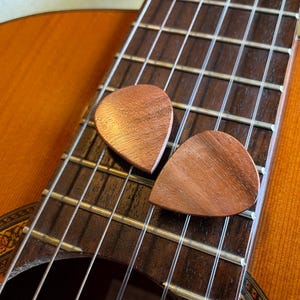 May include: Two wooden guitar picks rest on the fretboard of an acoustic guitar. The picks are a warm brown color, and the guitar has a light brown body. The strings and fretboard are visible.