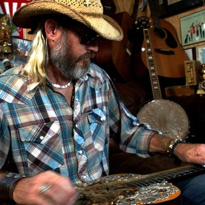 May include: A man wearing a straw cowboy hat and a plaid shirt plays a resonator guitar. The guitar has a silver and gold finish with a floral design. The man is sitting in a room with a variety of musical instruments in the background.