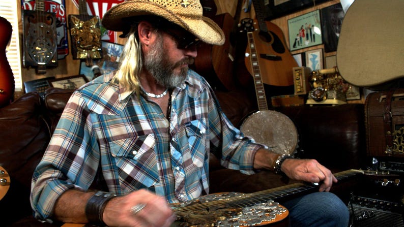 May include: A man wearing a straw cowboy hat and a plaid shirt plays a resonator guitar. The guitar has a silver and gold finish with a floral design. The man is sitting in a room with a variety of musical instruments in the background.