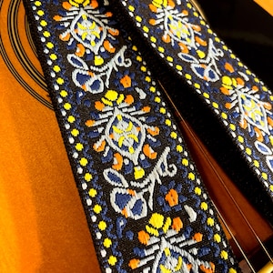 May include: Close-up of a guitar strap with a detailed floral pattern. The strap is black with a repeating design in yellow, orange, and blue. The strap is resting on the body of a wooden acoustic guitar.