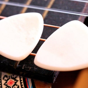 May include: Two off-white, triangular guitar picks rest on the fretboard of an acoustic guitar. The strings are visible, along with a portion of the guitar's body. The picks are smooth and appear to be made of a solid material.
