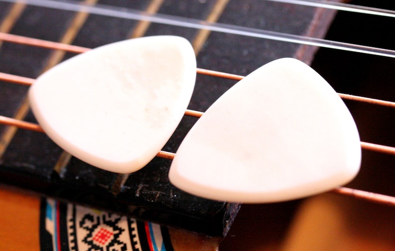 May include: Two off-white, triangular guitar picks rest on the fretboard of an acoustic guitar. The strings are visible, along with a portion of the guitar's body. The picks are smooth and appear to be made of a solid material.